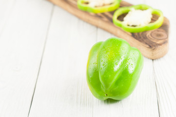 Whole green pepper beside row of slices of fresh ripe raw pepper on brown cutting board on old rural white wooden planks