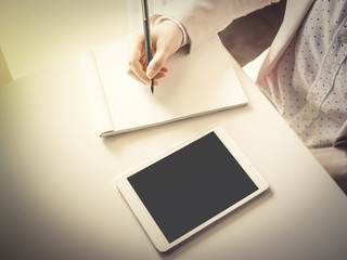 top view from right hand of business woman(30s to 40s) with pink or pastel suits check detail from tablet and writing on account book by black pen with soft focus background