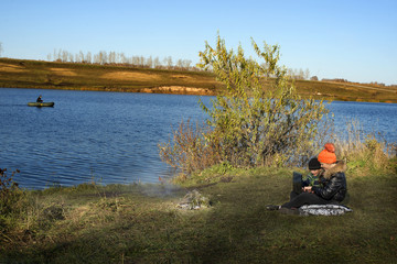 boy and girl sitting on the shore of a pond with a laptop in hands autumn day