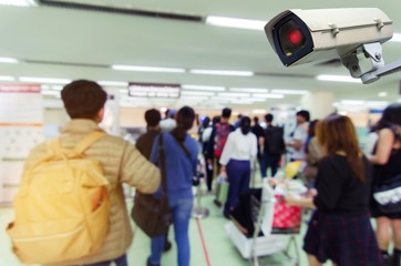 hand holding mobile phone and CCTV security indoor camera system operating with blurred image of people at immigration control at airport, internet, surveillance security, safety technology concept
