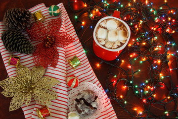 winter hot spicy drink cacao, with marshmallows and christmas chocolate cookies on background of striped blanket, pine cones, toys, decorations and electric lights for christmas tree with copy space
