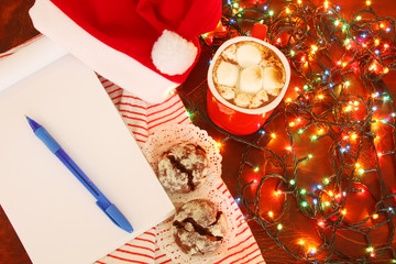 Santa's preparation for christmas presents delivery: hot spicy cacao with marshmallows, open notebook with list of names naughty and nice children, christmas chocolate cookies and christmas lights