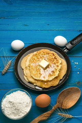 Hot delicious pancakes in frying pan on blue wooden table with flour and eggs. Pancake day background