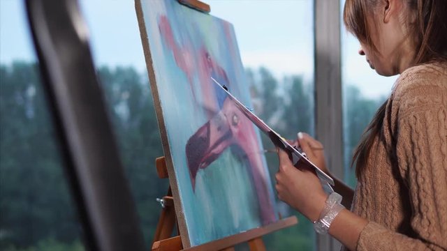 A young artist who holds a palette in her hand draws a picture of a flamingo on a blue background, in front are brushes stained with paint