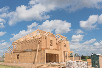 Wood frame house under construction and cloud blue sky in suburban Pearland, Texas, USA. New stick built 2 floor home wall covered by panels and sheathing. Portable toilet, pile of sand, gravel, logs