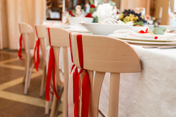 Wooden chairs with red tape stand in a row at a laid table. Rows of wooden chairs on decorated wedding ceremony. Chairs with red ribbons on for guests at the meeting.