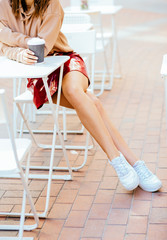 Vertical close up of woman's legs in red skirt and white sneakers holding disposable cup while sitting on street cafe with white tables and chairs.