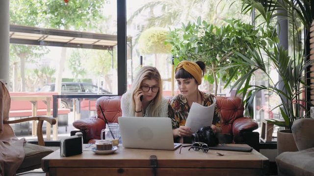 Young creatives, freelance workers, blonde and brunette pretty women, discuss and plan new project during meeting in trendy downtown cafe, have call on smartphone, point to laptop images