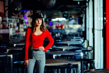 Provocated woman in red shirt and black peaked caps posing over blured dark loft pub or night club...
