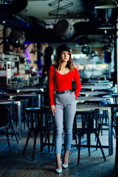 Vertical full height photo of fasionable provocated woman in red shirt and black peaked caps posing over blured dark loft pub or night club interior.