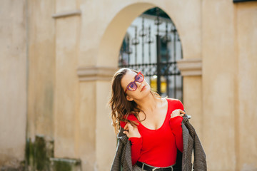 Charming fashionable woman in casual jacket in a cage and red shirt posing over old city building background. Weekend, travel and leisure concept.