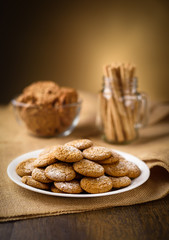 Honey ginger cookies in the foreground. Oatmeal raisin cookies and pirouette rolled wafers in the background.  Burlap on wooden table. Warm home atmosphere.