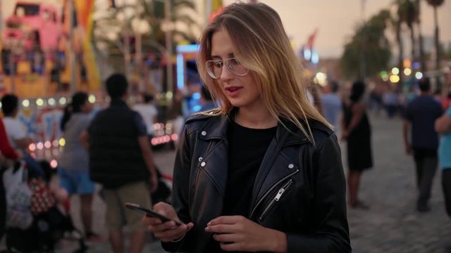 Cute shy, pretty and beautiful blonde woman or teenager in prescription glasses stands in middle of crowd on festival or carnival, chats and uses messaging or dating application on smartphone