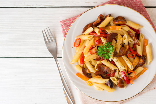 Penne pasta with mushrooms, bell pepper, chilli and parsley on white plate on white wooden background.