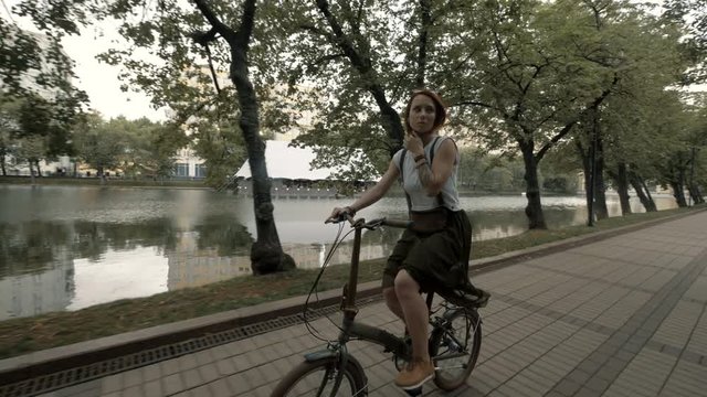Red haired woman riding a bicycle on background city lake. Woman bike city