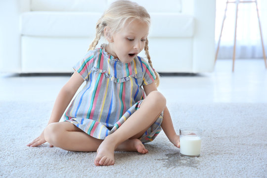 Cute little girl with glass of milk sitting on carpet near wet spot