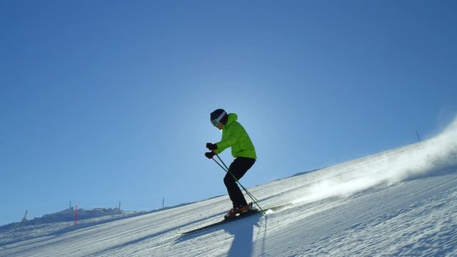 TRACKING SLOW MOTION: Alone recreational skier with bright  green jacket going over sun flare on a fresh morning groomed piste while skiing fast on cold perfect cloudless winter day at the ski resort.