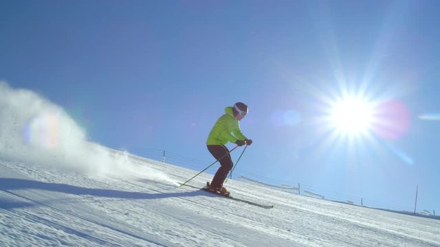 TRACKING SLOW MOTION: Young adult happy skier doing fast turns on a fresh winter morning groomed piste at European ski resort in the Alps during cloudless day. Sun flare shinning in the camera.
