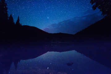 Mountain lake Synevir at night with starry sky and reflections in the water. Natural outddors travel dark background. Carpathian, Ukraine