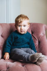 Vertical portrait of innocent little child with blue eyeys and plump cheeks, looks directly into camera, wait for children as has birthday, sits in comfortable pink armchair in living room. Children