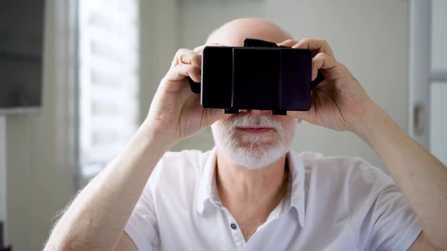 Good-looking handsome senior man in white using VR 360 glasses at home. Active modern elderly people concept. Seeing something unusual and exciting, learning to use contemporary technologies.