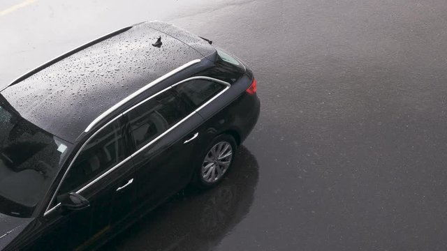 Rain Falls On The Already Rain Speckled Rooftop Of A Black Car Parked On Black Asphalt. High Angle, Zoom In.