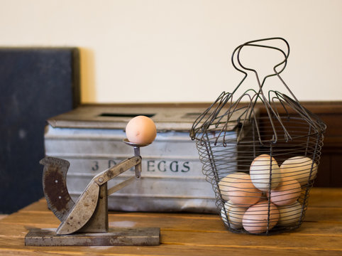 A vintage Egg Grader to determine the size of a hen's egg. Additional eggs in a wire basket and a metal container for eggs are in the background.