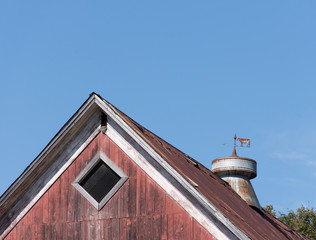 Close up of a weathered, rusty vintage weather vane that shows an arrow and a cow. It is atop an old red barn and against blue sky.