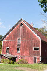 Obraz premium Old Red Barn in disrepair with a cow weather vane on its roof. Blue sky above barn provides copy space.
