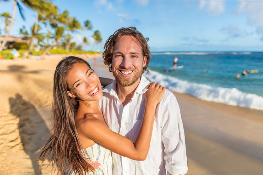 Couple in love hugging happy together on beach travel holidays at sunset. Beautiful Asian woman and caucasian man portrait. Young couple lovers relaxing on honeymoon.