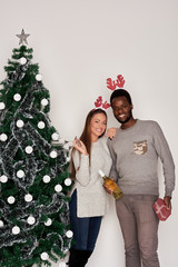 Multi ethnic couple posing with Christmas gift, champagne and wine glasses in hands. Christmas tree next to them / Christmas