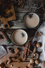 Fototapeta na wymiar spiced drink with milk in metal mugs with cinnamon sticks, anise, chocolate slices, assorted nuts, vintage pot, textile on rustic wooden table