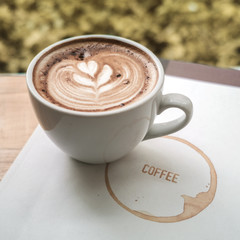 A cup of coffee latte art with heart in a white cup on a book of coffee with green leave frame