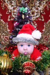 Newborn naughty Santa baby with amazing blue eyes dressed  in red santa hat and jacket shows tongue out laying on tinsel and christmas decorated background, Christmas tree behind of him. New year 2018