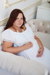 Peaceful beautiful pregnant woman with happy smile in  white dress sitting on white sofa with hand on belly.