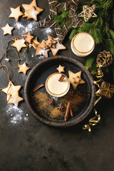Eggnog Christmas milk cocktail with cinnamon, served in two glasses with shortbread star shape sugar cookies different size, decor toys, fir branch over dark texture background. Top view with space