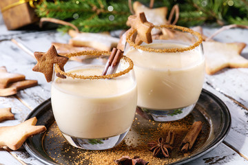 Cocktail de lait de Noël au lait de poule à la cannelle, servi dans deux verres sur un plateau vintage avec des biscuits sablés en forme d& 39 étoile, des jouets de décoration, une branche de sapin sur une table en bois blanc. Fermer