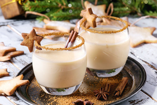 Eggnog Christmas milk cocktail with cinnamon, served in two glasses on vintage tray with shortbread star shape sugar cookies, decor toys, fir branch over white wooden plank table. Close up