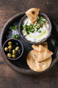 labneh middle eastern lebanese cream cheese dip with olive oil, salt, herbs served with olives, traditional pita bread on terracotta plate over dark texture wooden background. Top view with space