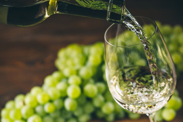 Pouring white wine into a glass with a bunch of green grapes against wooden background