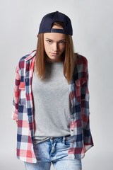 Angry teen girl wearing checkered shirt and baseball cap over grey background looking at camera sullen, front view