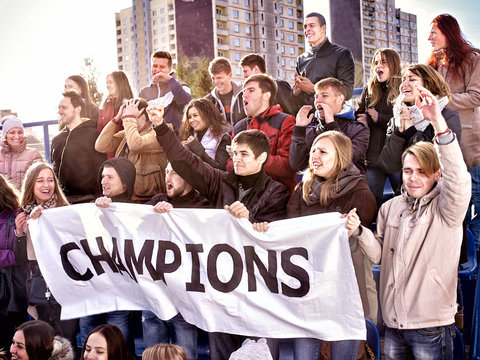 Fans Cheering In Stadium Holding Champion Banner And Singing On Tribunes. Large Group Young People Together Support Your Favorite Team. Cold Weather Outdoor. Beginning Of Football Match.