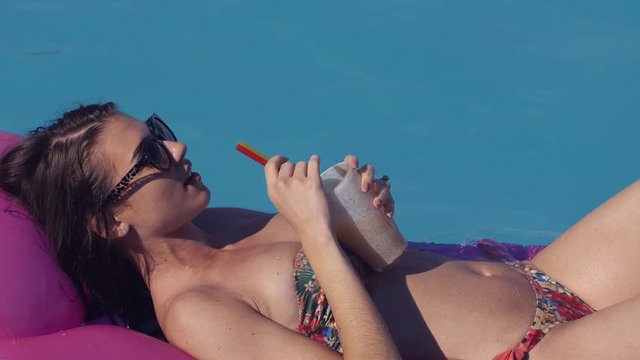 Beautiful young woman relaxing in swimming pool in summer, lying on inflatable Lounge in sunglases Summer time. She drinks a cocktail