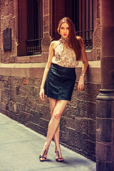 Young European Woman with long hair, traveling in New York, wearing light patterned, sleeveless deep v neck top, black leather skirt, sandals, standing against vintage wall with windows on street..