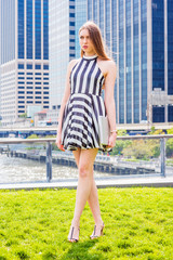 European college student traveling, studying in New York, wearing black and white striped short dress, sandals, holding laptop computer, standing on green lawn in business district, looking forward..