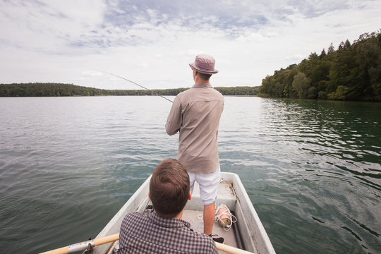 A man is rowing a rowboat while his friend is fly fishing from the boat on a  lake.
