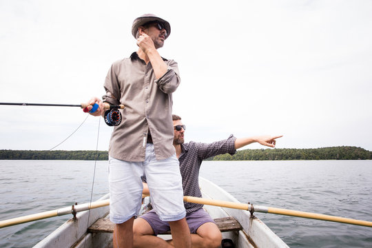 A man is rowing a rowboat while his friend is fly fishing from the boat on a  lake.