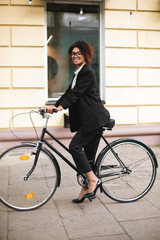 Young smiling African American girl in glasses standing with bicycle and happily looking in camera. Beautiful lady with dark curly hair in suit cycling along the street