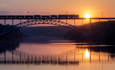 freight train rides on the railway bridge over the river during sunse