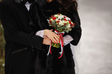Winter wedding. Bride and groom in black fur coat holding red bouquet of fresh flowers